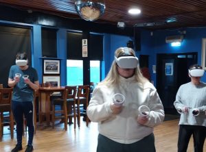 Three students are standing in a room wearing a VR headset and holding VR controllers.
