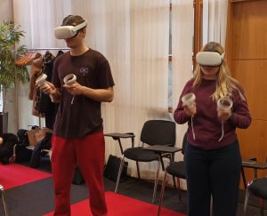 Two students standing upright with VR headsets on and holding VR controllers in their hands.